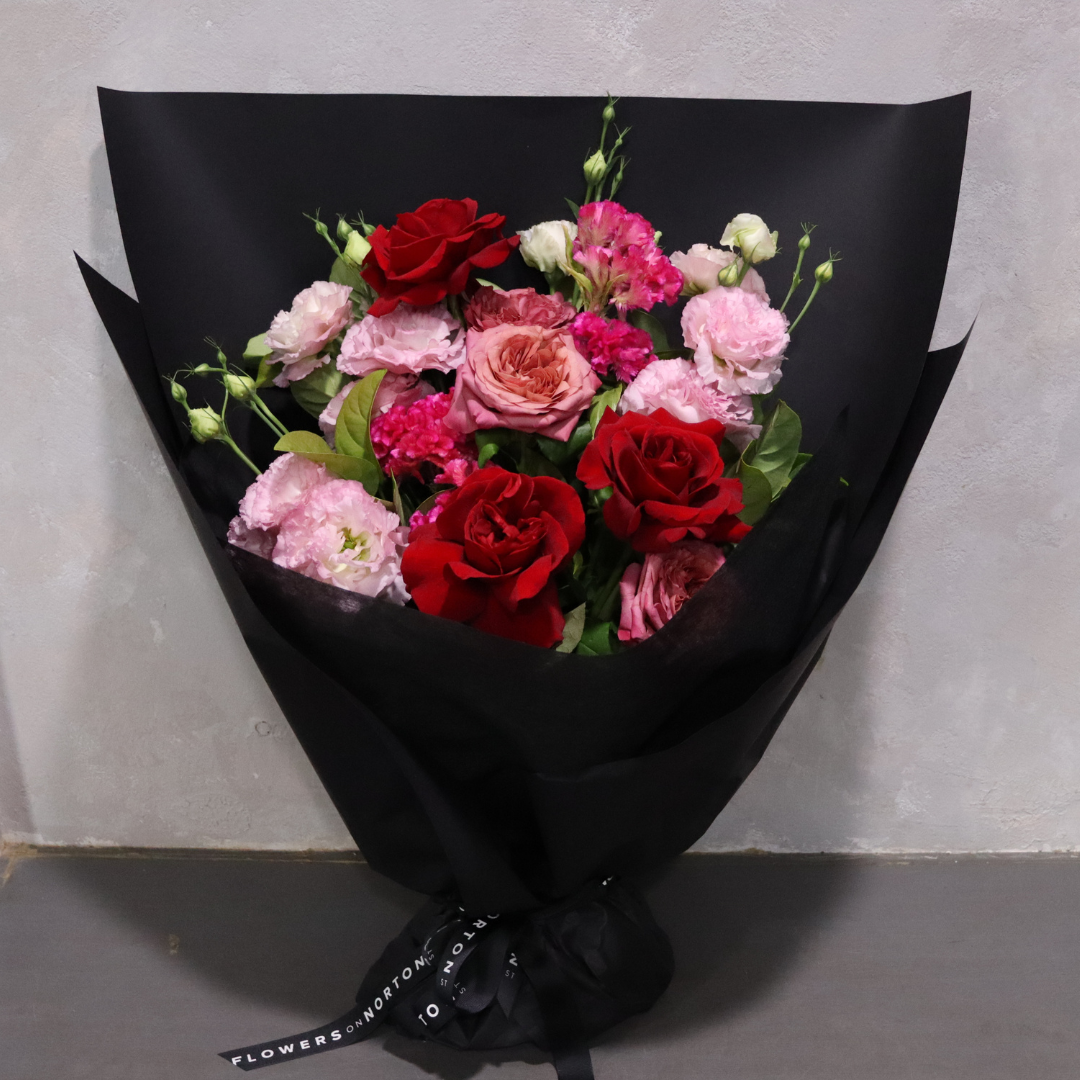 Introducing the Sweet Romance bouquet from Flowers on Norton St: a breathtaking arrangement wrapped in black paper, showcasing fresh red roses, pink peonies, and lush green leaves. This vibrant ensemble offers an elegant contrast against its neutral backdrop.