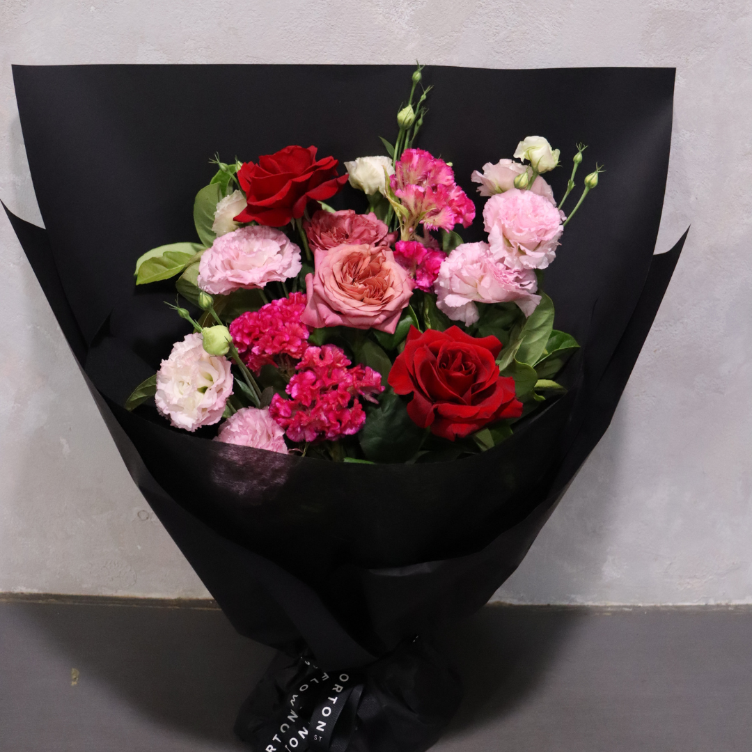 Introducing the Sweet Romance by Flowers on Norton St: a sophisticated arrangement of bright red and gentle pink roses and carnations, complemented by lush greenery. Presented in elegant black wrapping paper and adorned with a ribbon, it's the perfect highlight of our Valentine's Day Collection against a neutral backdrop.