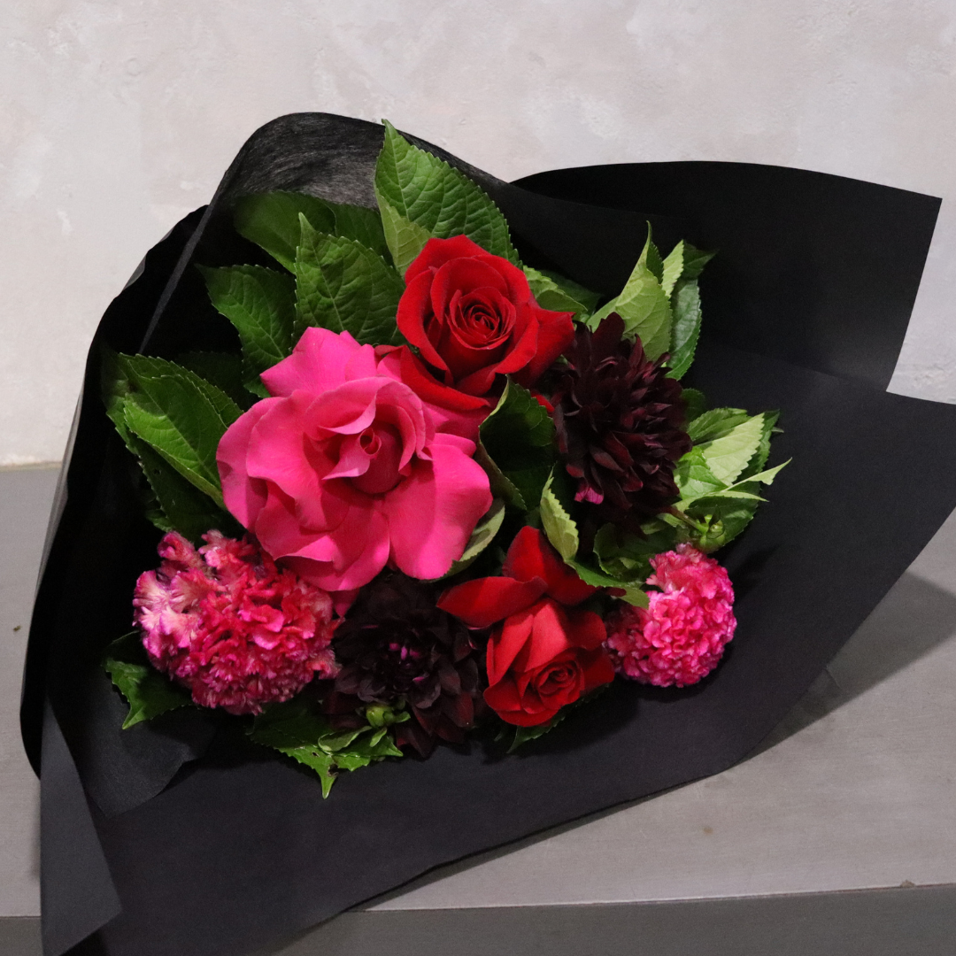 The "With Love" bouquet from Flowers on Norton St, wrapped in elegant black paper, includes a stunning combination of pink and red roses, dark burgundy flowers, and pink carnations complemented by lush green leaves. This delightful arrangement is beautifully presented on a gray surface.