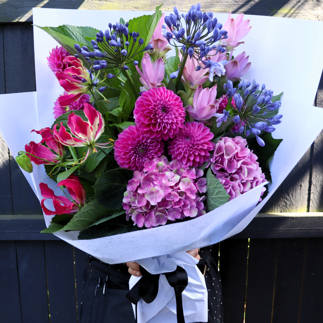 The "Fireworks" bouquet from Flowers on Norton St features a vibrant array of pink and purple flowers, including dahlias, hydrangeas, agapanthus, and gloriosa. It is elegantly wrapped in white paper with a black ribbon, offering a striking contrast against a dark background.