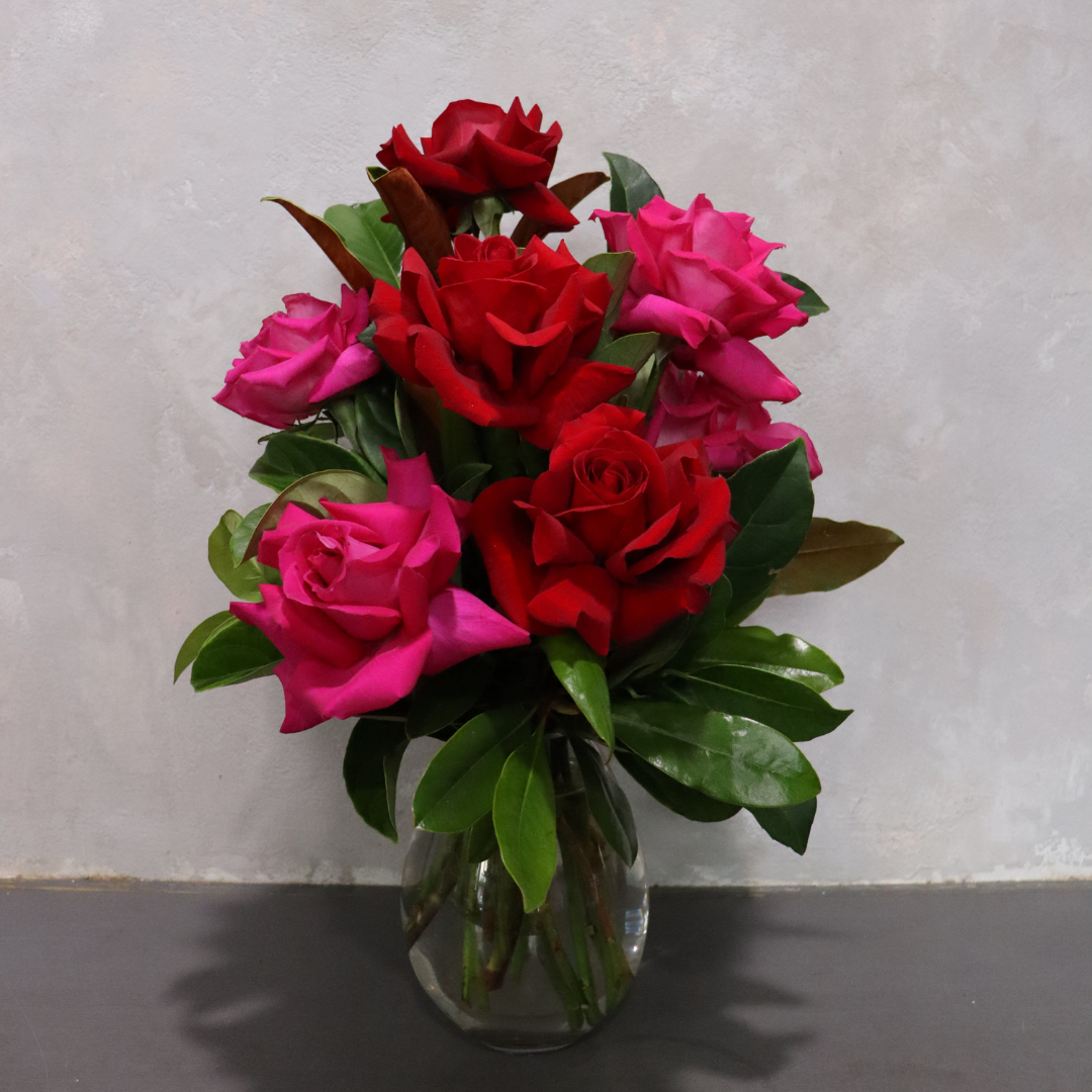 A romantic bouquet from Flowers on Norton St, the Be Mine Rose Bouquet overflows with vibrant red and pink roses, complemented by lush green leaves against a light gray textured background.