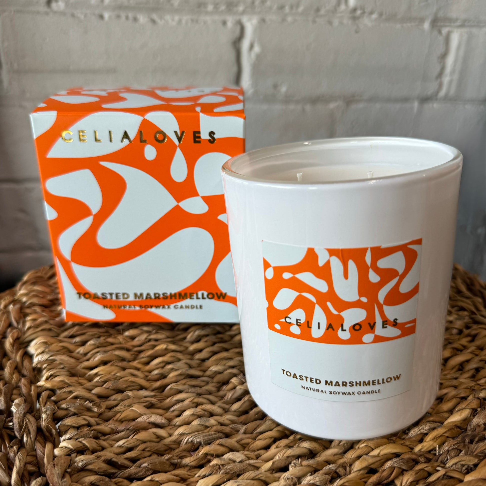 A hand-poured white candle labeled "Toasted Marshmallow," adorned with orange abstract patterns, rests on a woven surface beside its matching box. The brand name "Flowers on Norton St" is visible on both, highlighting distinctive fragrances against a white brick wall.