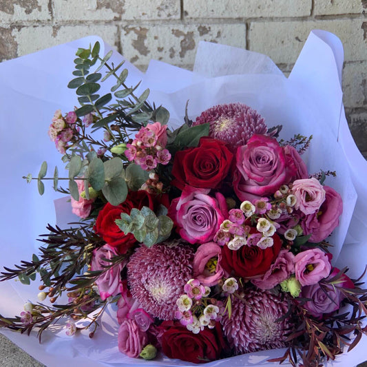 Introducing the Berry Bliss Purple Bouquet by Flowers on Norton St: a vibrant collection of red and pink roses, complemented with pink chrysanthemums and delicate small white flowers. Accented with lush green eucalyptus leaves, this beautiful arrangement is elegantly wrapped in white paper and displayed against a rustic brick background.