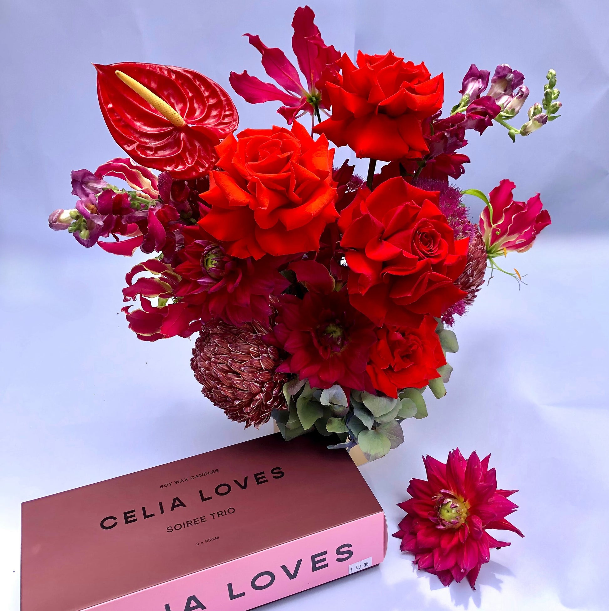 A vibrant red and burgundy flower arrangement featuring roses and anthuriums from the "A Soiree Affair Red Bouquet" by Flowers on Norton St adorns a vase next to a pink box labeled "Celia Loves." A single red flower rests alongside, completing the elegant scene reminiscent of this exquisite gift set.