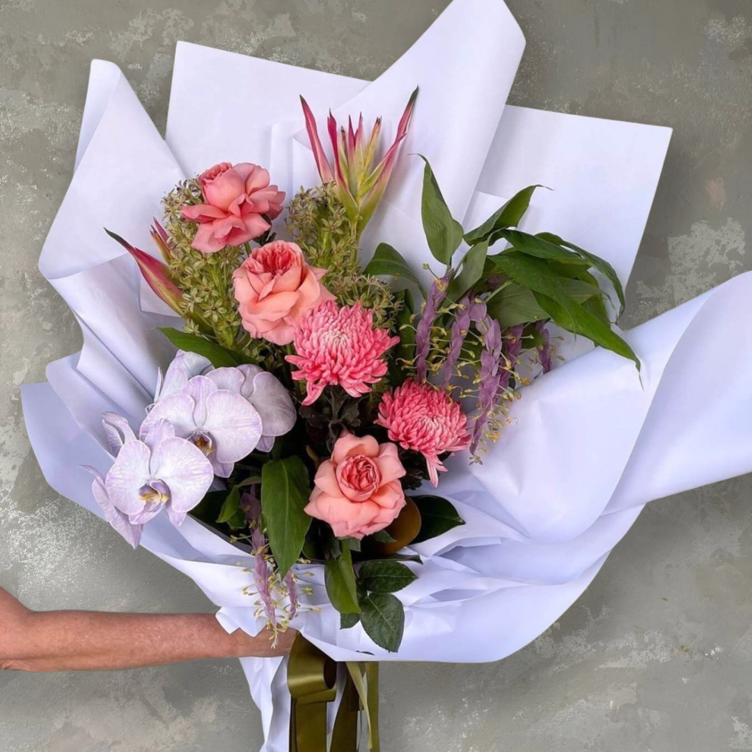 A person holds a Floral Expression bouquet from FlowersonNortonSt, featuring pretty pinks and greens with chrysanthemums, tropical flowers, and white orchids against a gray background.