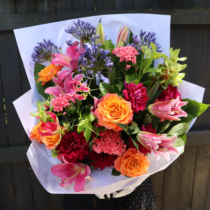 Introducing the "Summer Harvest" by FlowersonNortonSt—a stunning arrangement of vibrant luxury flowers. This bouquet showcases pink and orange roses, pink lilies, purple agapanthus, and red dahlias wrapped in elegant white paper against a dark wooden backdrop. It beautifully captures the essence of bright colored flowers with sophistication.