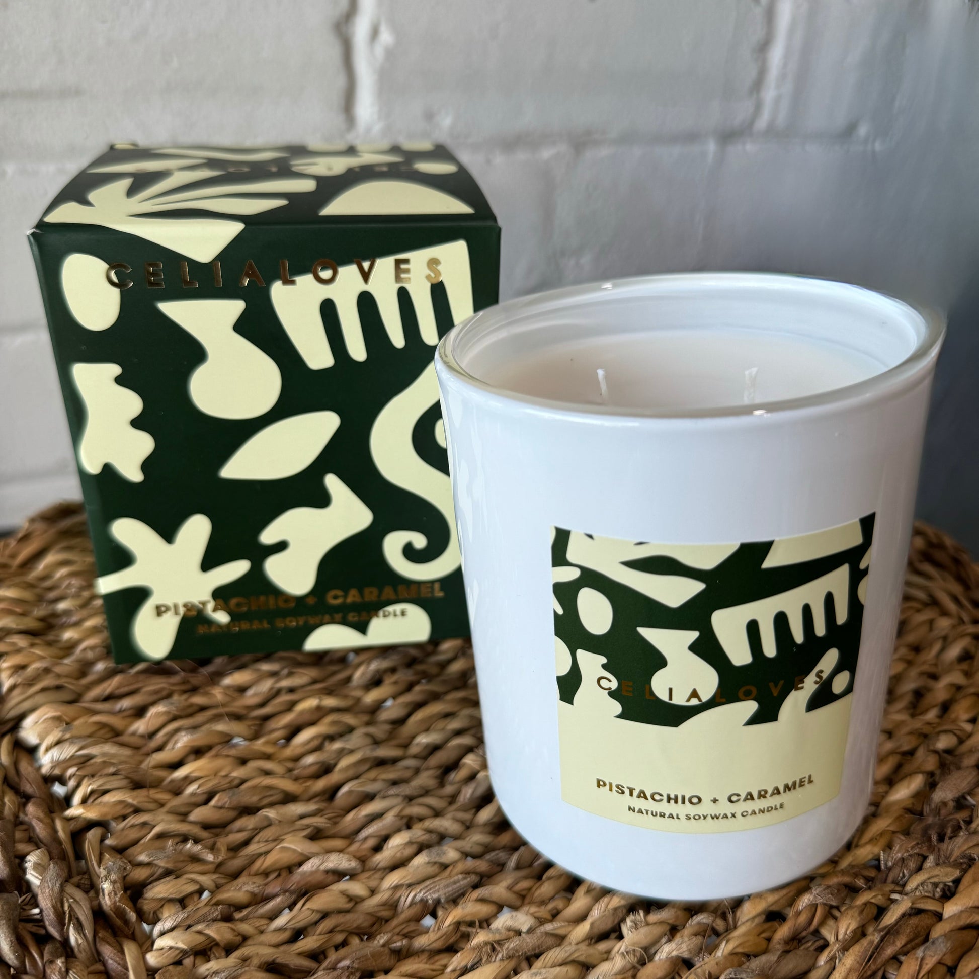 A soy candle jar in white, adorned with abstract green and beige patterns and labeled "Pistachio + Caramel," delivers unique fragrances from Flowers on Norton St’s Celia Loves Candles. It rests on a woven surface beside its hand-poured counterpart, housed in an equally patterned box that prominently features "CELIA LOVES.