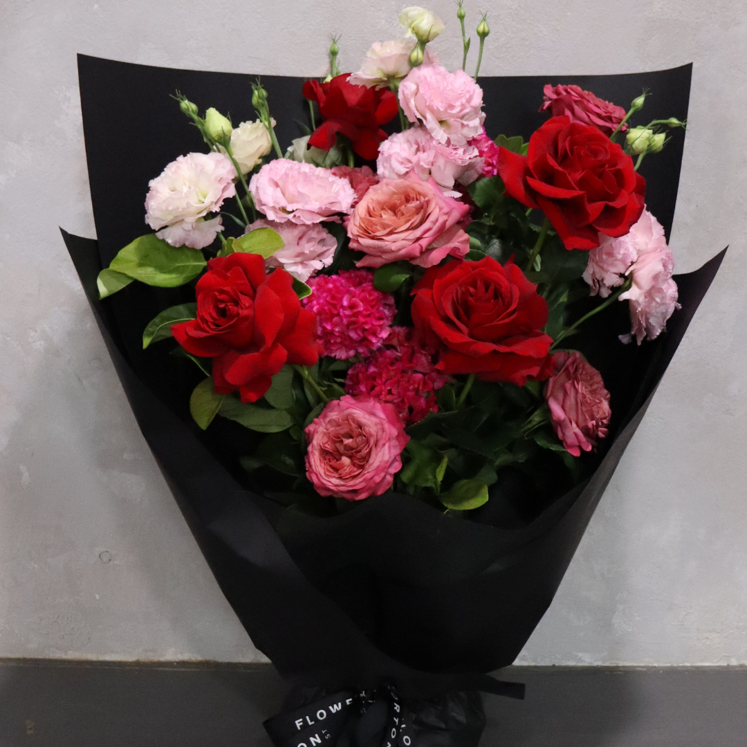 The Sweet Romance floral arrangement by Flowers on Norton St showcases an elegant blend of red and pink roses, complemented by pink carnations and green leaves. It is artfully wrapped in chic black paper, making it a perfect addition to the Valentine's Day Collection against a light gray background.