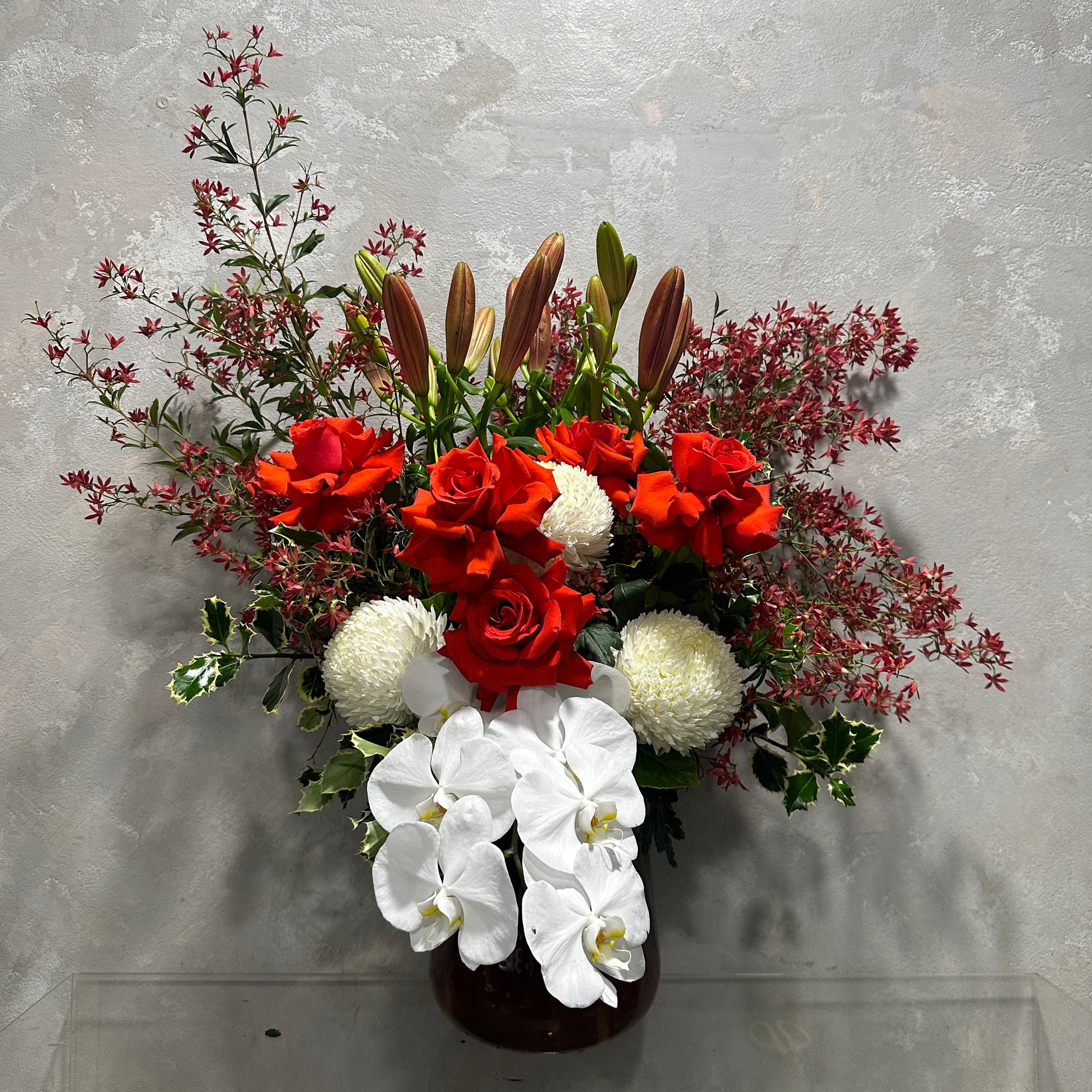 The "Aussie Christmas Vase" by Flowers on Norton St beautifully captures the essence of holiday cheer with its floral arrangement of red roses, white orchids, and white chrysanthemums, complemented by Australian native Christmas bush and greenery. The dark leaves and small red branches are elegantly displayed in a dark vase against a light gray textured wall.