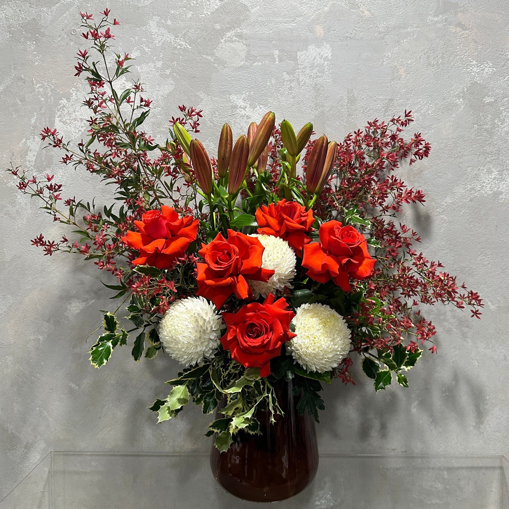 The Aussie Christmas Vase by Flowers on Norton St showcases a stunning floral arrangement in a dark vase, featuring red roses, white chrysanthemums, budding lilies, and vibrant Australian native Christmas bush foliage. Positioned against a textured gray wall, it’s an ideal piece for bringing cheer to the holiday season.