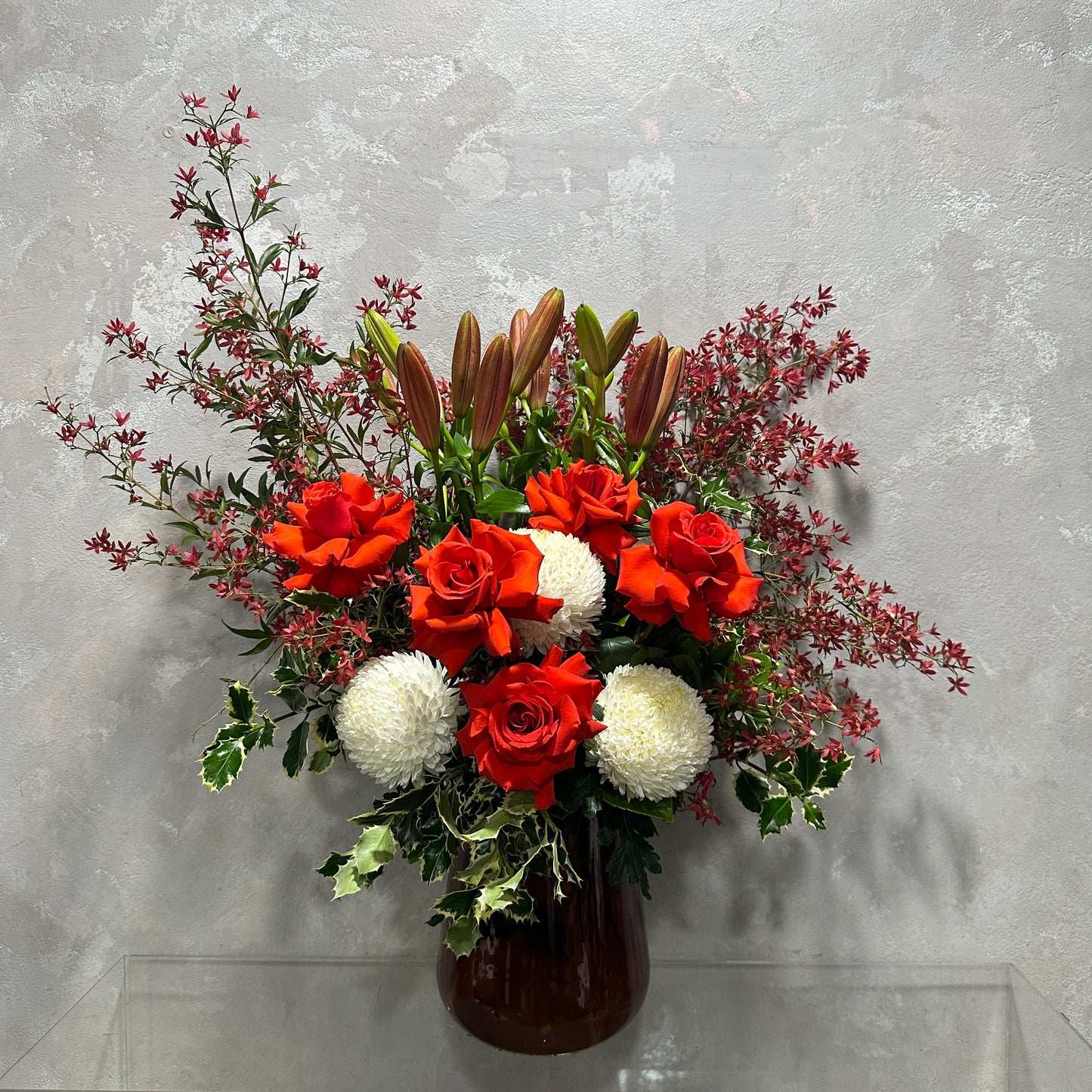 The Aussie Christmas Vase from Flowers on Norton St, showcasing vibrant red roses, white chrysanthemums, pink lilies, and lush greenery, adds festive holiday charm to a textured gray wall.