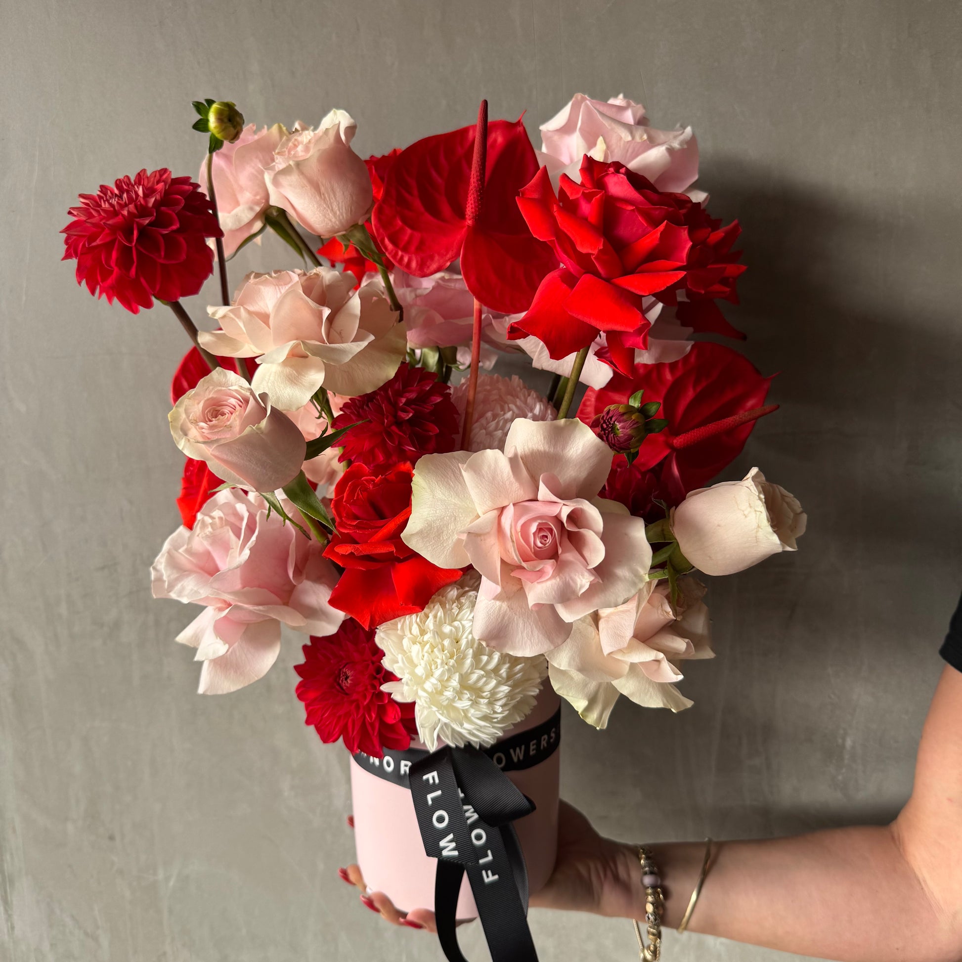 A hand displays the Be My Valentine Vase Arrangement by Flowers on Norton St, featuring Colombian red and pink roses, dahlias, anthuriums, and more in a pink pot with a black ribbon against a gray background—perfect for Valentine's Day.