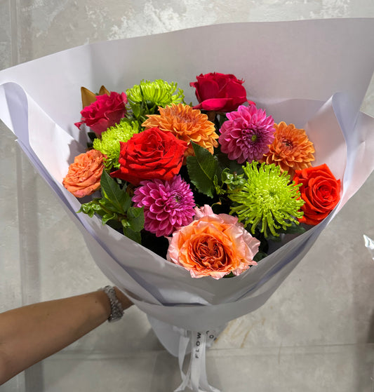 The Bigger Pop of Colour Mixed Flower Bouquet from FlowersonNortonSt features a vibrant mix of seasonal favorites: red roses, orange and pink dahlias, green spider chrysanthemums, and orange roses. Wrapped in white paper, it shines beautifully against a light gray background.