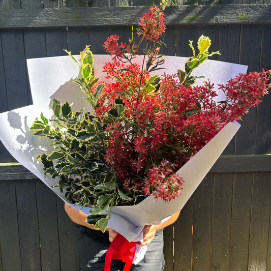 A person holds Flowers on Norton St's "Christmas Joy Bouquet," featuring red flowers and variegated green leaves, wrapped in white paper with a red ribbon. Holly sprigs evoke Christmas vibes against a dark wooden fence backdrop.