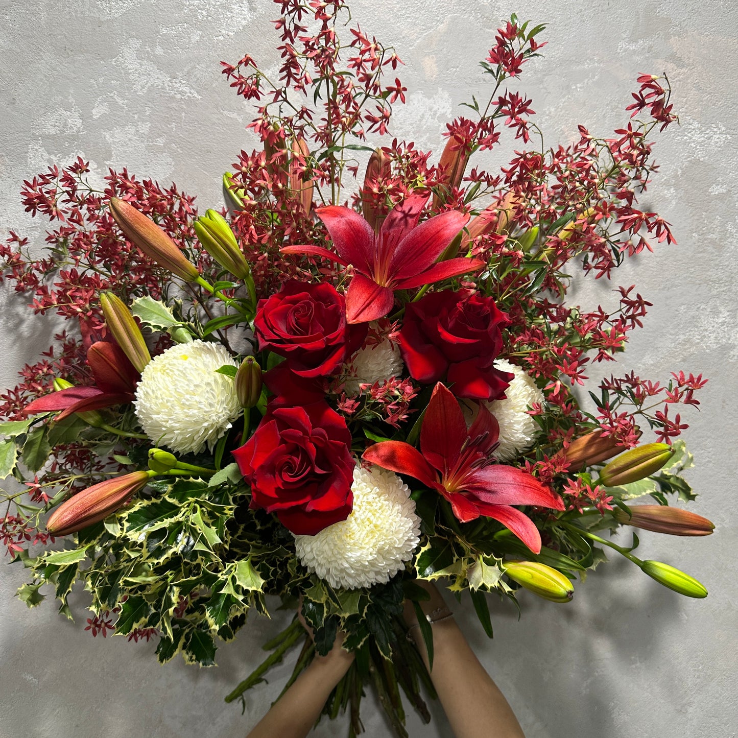 The Christmas Lily + Rose Bouquet by Flowers on Norton St features a lively mix of red roses, oriental lilies, and white chrysanthemums complemented by lush greenery against a textured gray background, embodying the spirit of holiday decor. The bouquet is gently cradled in hands.