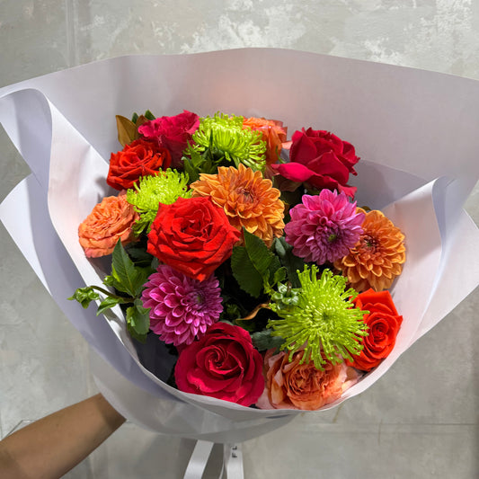 The Even Bigger Pop of Colour Mixed Bouquet by FlowersonNortonSt features vibrant red, orange, and pink roses, dahlias in orange and pink, and lively green chrysanthemums. Elegantly wrapped in white paper against a neutral background, it's perfect for expressing gratitude or congratulating someone special.