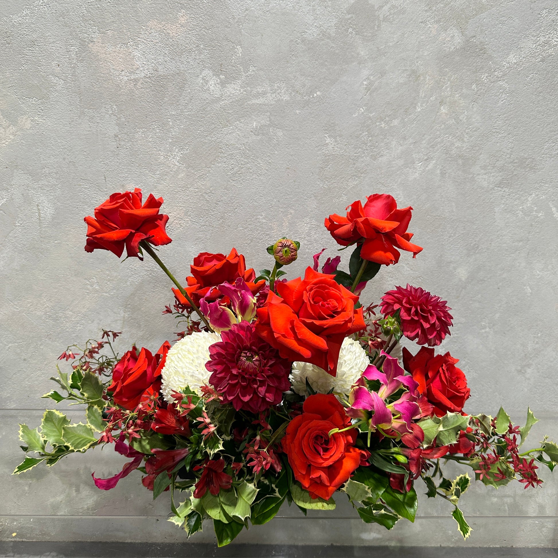 Experience a touch of natural elegance and charm with the "Festive Floral Table Centre" from Flowers on Norton St. This vibrant centerpiece showcases red roses, dark pink dahlias, and white chrysanthemums, beautifully accented by green foliage against a textured gray background, making it an ideal choice for enhancing your holiday table decor.