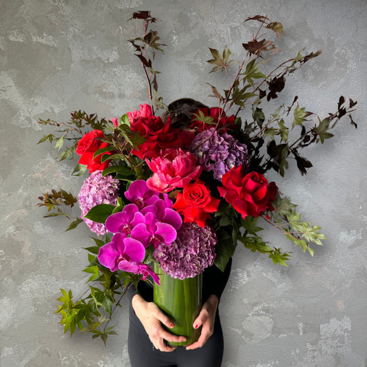 An individual cradles The Floral Cha-Cha Vase Arrangement by Flowers on Norton St, featuring red roses, purple hydrangeas, and pink orchids against a textured gray backdrop. Green and brown foliage adds contrast while the bouquet mostly hides the person's face.