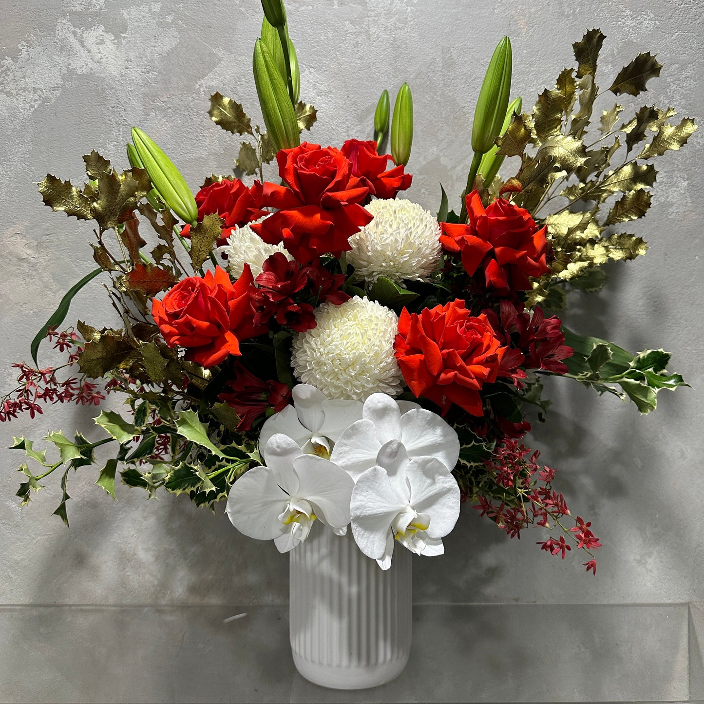 Celebrate the holiday season with a vibrant floral arrangement featuring red roses, white chrysanthemums, and white orchids elegantly displayed in the Gold Holly Xmas Vase by Flowers on Norton St. This stunning bouquet is complemented with lush green leaves and lily buds, set against a textured gray backdrop, making it an ideal choice for festive decor.