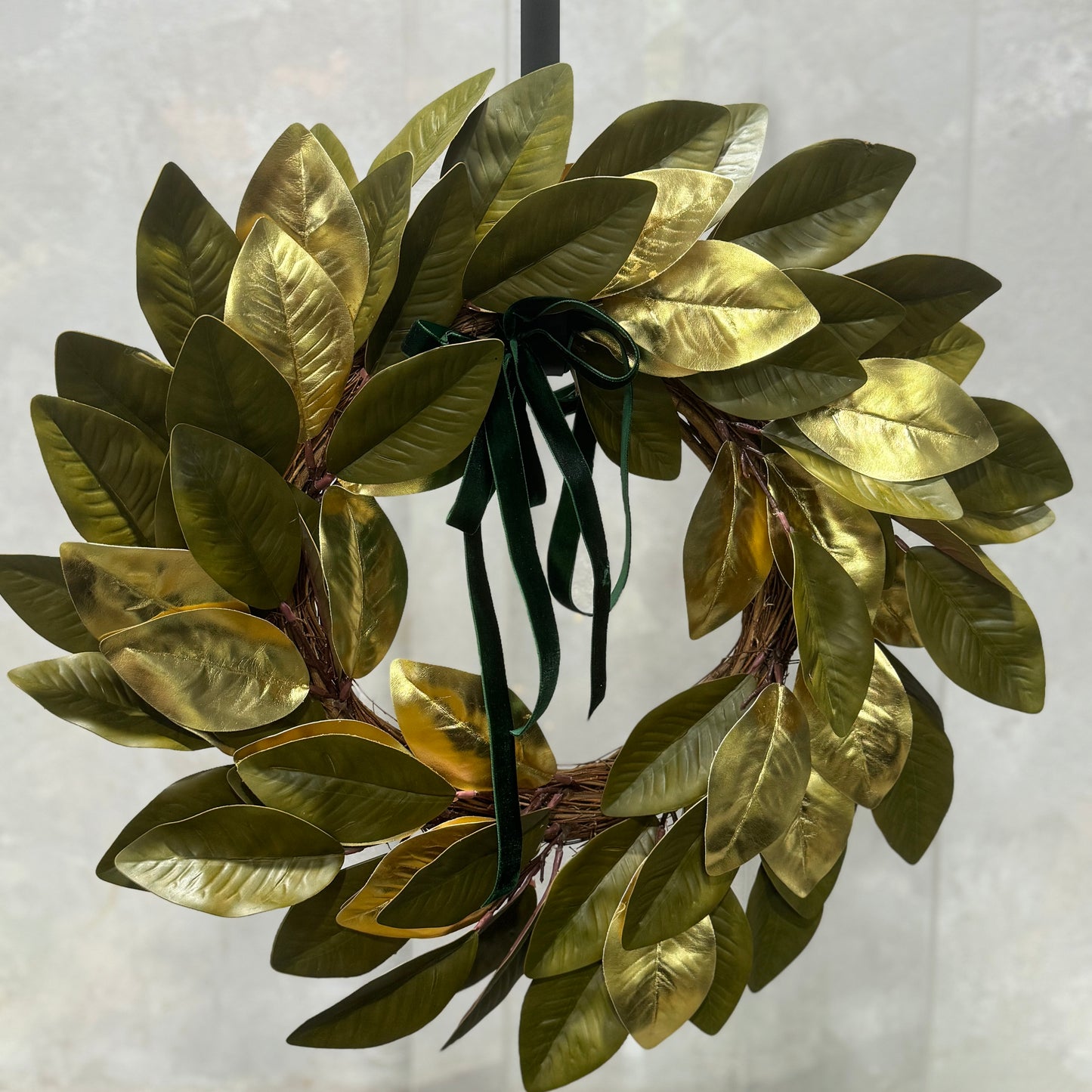 The Golden Glow Wreath by Flowers on Norton St, adorned with shiny gold and green leaves, is elegantly suspended against a light, textured background. It showcases festive red poinsettias and is topped with a dark green ribbon bow.