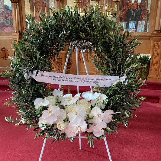 A charming Peace Wreath with Feature Spray, offered by FlowersonNortonSt, adorned with green leaves and white flowers, is displayed on an easel in the church. A ribbon draped across the wreath says, "To the greatest dad: You will always be in our hearts forever. Rest In Peace love your wife and children.