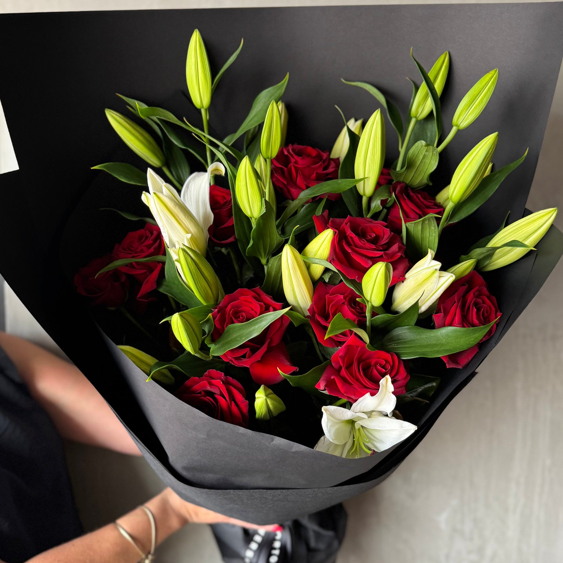 The White Lily + Red Rose Bouquet by Flowers on Norton St showcases vibrant red roses and partially blooming white lilies, elegantly wrapped in black paper. Perfect for anniversary or Valentine's Day, it looks stunning when held by someone with a silver bracelet against a neutral background.