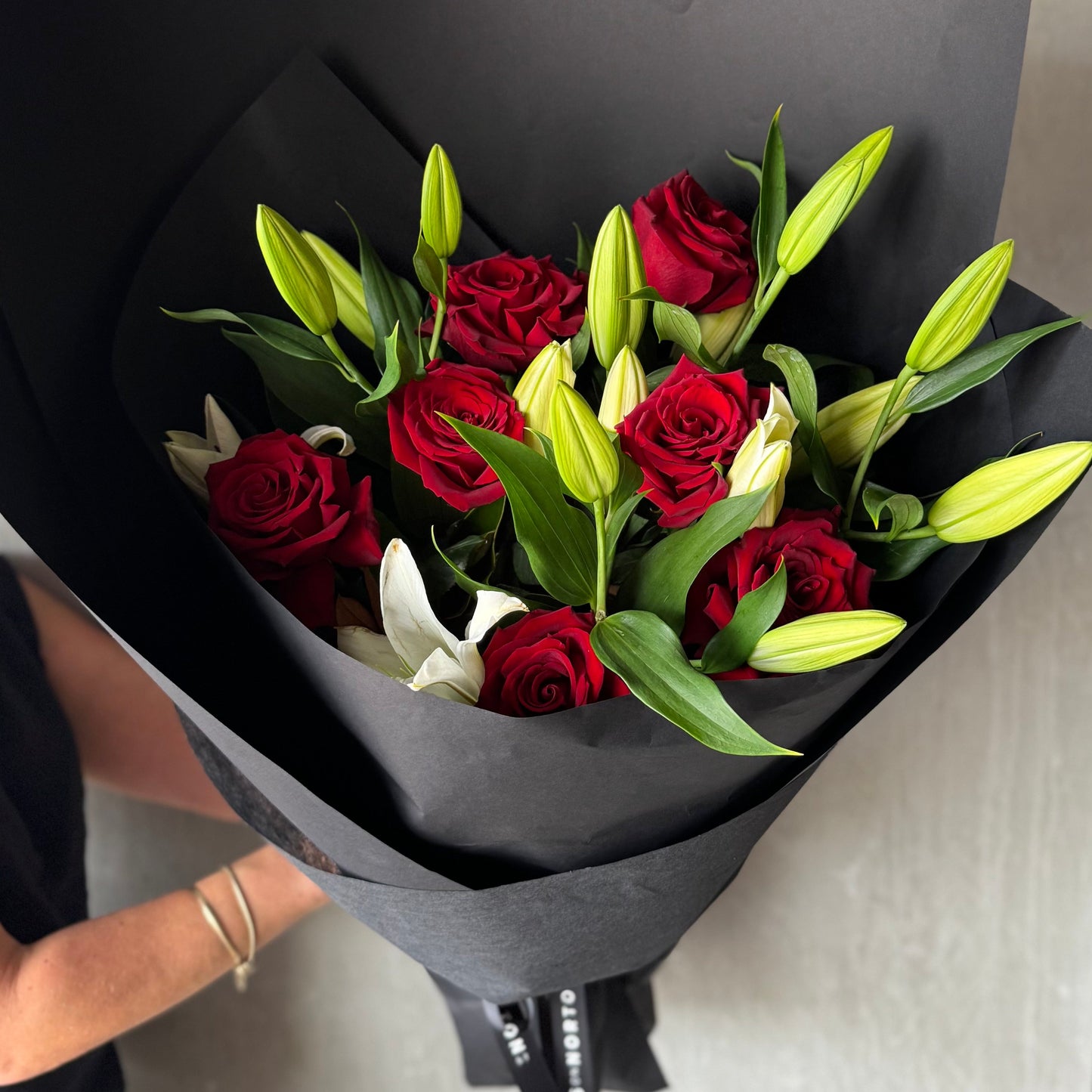 The Flowers on Norton St White Lily + Red Rose Bouquet, wrapped in black paper and held by a person with a silver bracelet, makes the perfect Valentine's Day gift.