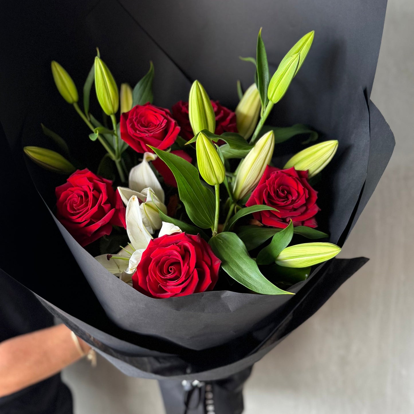 A person elegantly holds a Flowers on Norton St White Lily + Red Rose Bouquet wrapped in black paper, perfect for Valentine's Day.