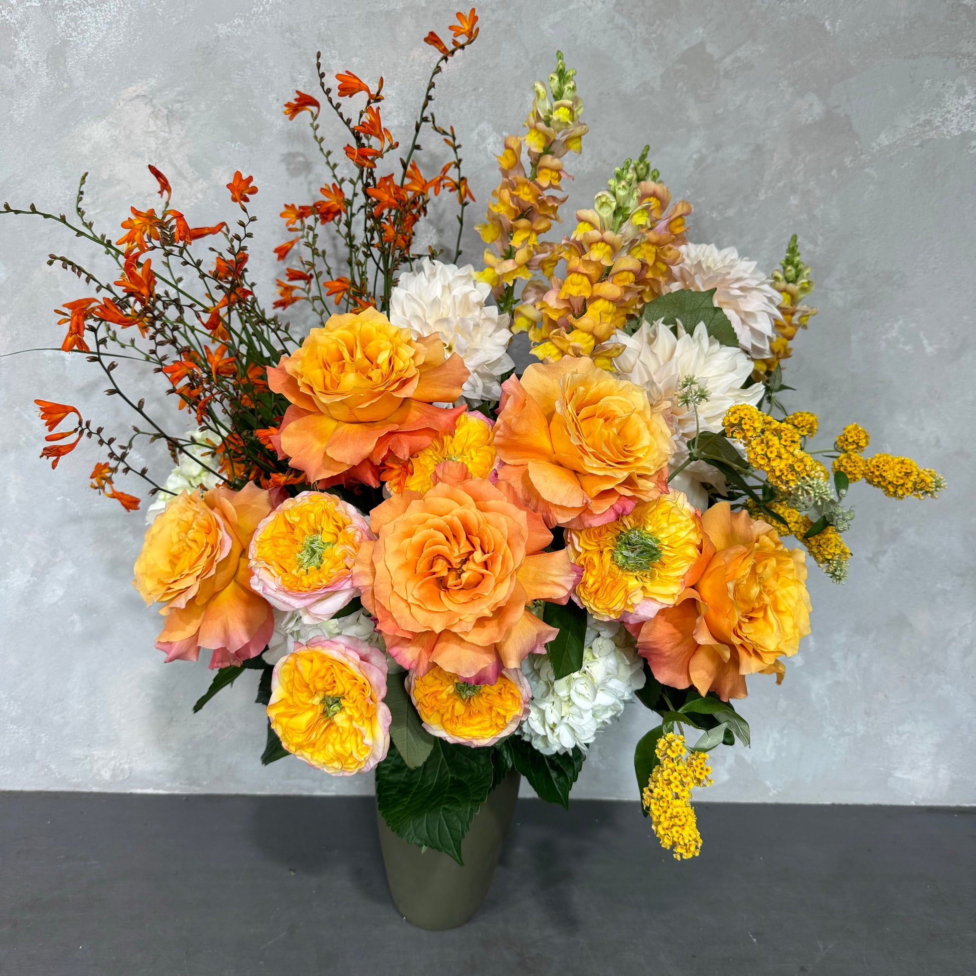 A vibrant floral arrangement in the Sweet like a Sunrise Vase from Flowers on Norton St showcases orange roses, white hydrangeas, and bright yellow snapdragons. It is beautifully accented with orange crocosmia and yellow solidago set against a light gray background, creating a cheerful atmosphere.