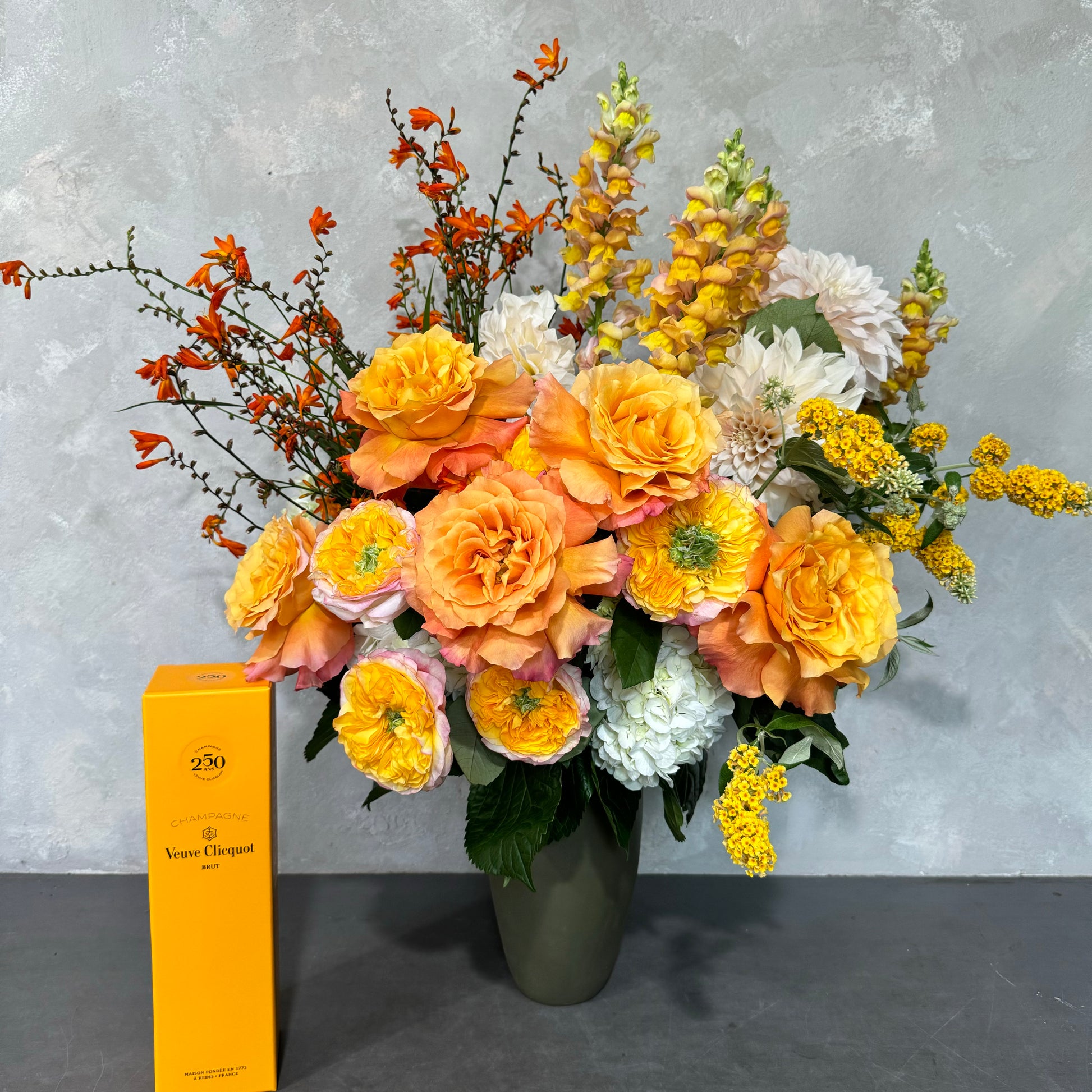 The Sweet like a Sunrise Vase by Flowers on Norton St showcases a delightful floral arrangement with golden blooms of orange and yellow roses, complemented by white flowers and lush green foliage. Beside it sits a yellow box of Veuve Clicquot champagne, all set against a light gray background for an uplifting ambiance.