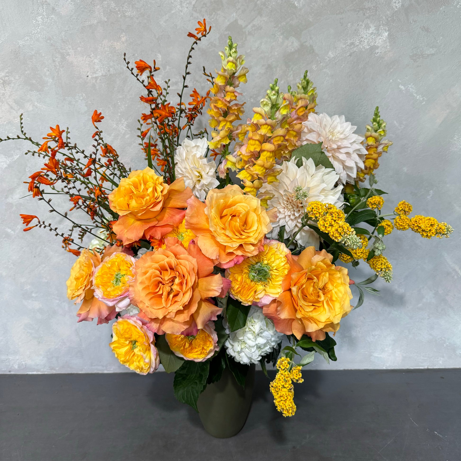 A lively floral display showcasing the golden hues of orange and yellow roses, complemented by yellow snapdragons, white dahlias, and delicate sprigs of small orange and yellow flowers, elegantly arranged in the Sweet like a Sunrise Vase from Flowers on Norton St against a subtly textured light gray background.