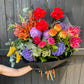 Introducing "My Favourite Colour is Rainbow 🌈" by FlowersonNortonSt, a vivacious bouquet showcasing red roses, orange snapdragons, purple hydrangeas, pink dahlias, and yellow blooms all elegantly wrapped in black paper. A colorful ribbon ties together this festive creation, making it the perfect choice for pride season. Displayed beautifully against a dark wooden fence backdrop.