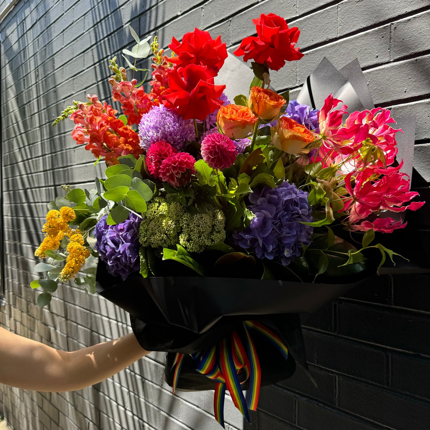 A vibrant bouquet titled "My favourite colour is Rainbow 🌈" by FlowersonNortonSt, featuring red, pink, purple, and orange flowers with green foliage. The arrangement is elegantly wrapped in black paper and secured with a colorful striped ribbon. It's beautifully displayed against a textured gray brick wall, capturing the essence of pride season.