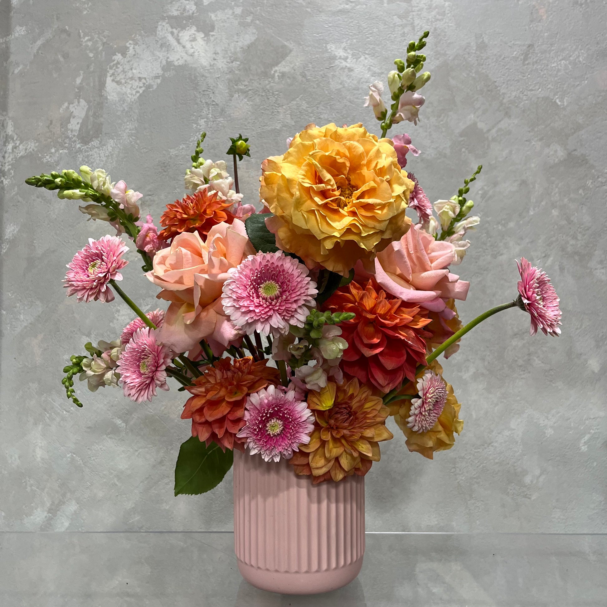 The Fairyfloss by Flowers on Norton St presents a breathtaking array of vibrant yellow, orange, and pink roses and dahlias in a charming pink vase. The lush green leaves contrast elegantly with the textured gray backdrop, creating an enchanting visual display in this exquisite ceramic pot creation.