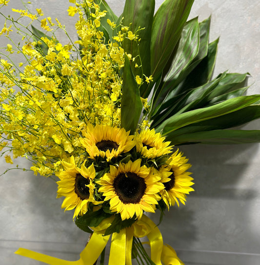 Introducing the Radiant Rays Bouquet by Flowers on Norton St: a delightful display featuring vibrant yellow sunflowers and yellow dancing lady orchids, accentuated by their dark centers and surrounded by large green leaves. The arrangement is cheerfully adorned with a bright yellow ribbon against a soft gray background.