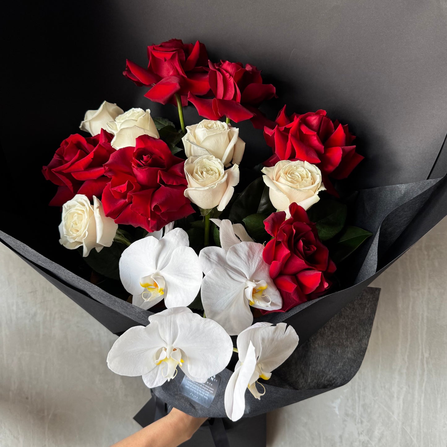 The Red and White Bud + Bloom Rose Bouquet by Flowers on Norton St features roses and white orchids elegantly wrapped in black paper. This 12-rose bouquet is beautifully arranged on a light gray surface, embodying classic charm.