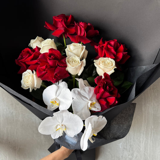 The Bud + Bloom Rose Bouquet with Phalaenopsis Orchid by Flowers on Norton St showcases red and white roses elegantly arranged with orchids, wrapped in black paper. A hand holds this exquisite bouquet against a neutral, textured background.