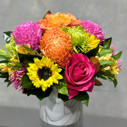 The Sunny + Bright Arrangement by Flowers on Norton St presents a vibrant floral display in a ceramic pot, showcasing pink roses, orange and pink chrysanthemums, green button poms, and yellow sunflowers against a soft gray background for a colorful bouquet.