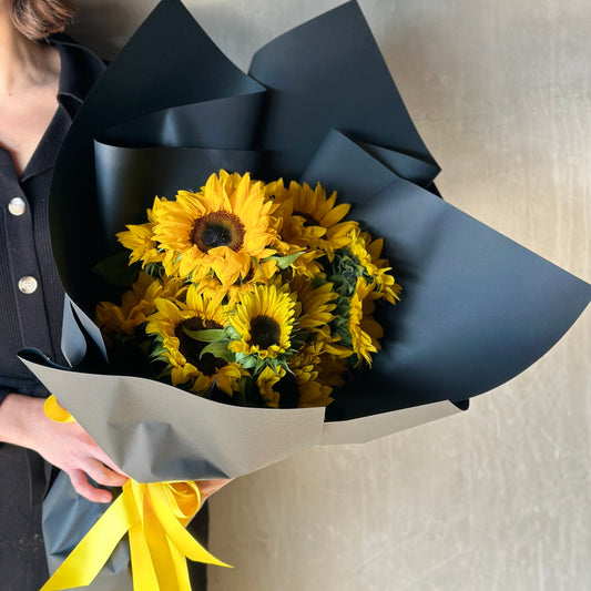 A person holds the breathtaking Sungold Surprise bouquet from Flowers on Norton St, featuring a burst of vibrant yellow sunflowers elegantly wrapped in black paper and tied with a matching yellow ribbon, perfect for a special celebration. They're standing against a plain textured background.