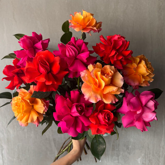 The Rose Trio Bouquet by FlowersonNortonSt features a vibrant blend of multicolored, handcrafted roses in shades of red, pink, and orange. These full-bloom roses are set against a gray background and framed by lush green leaves.