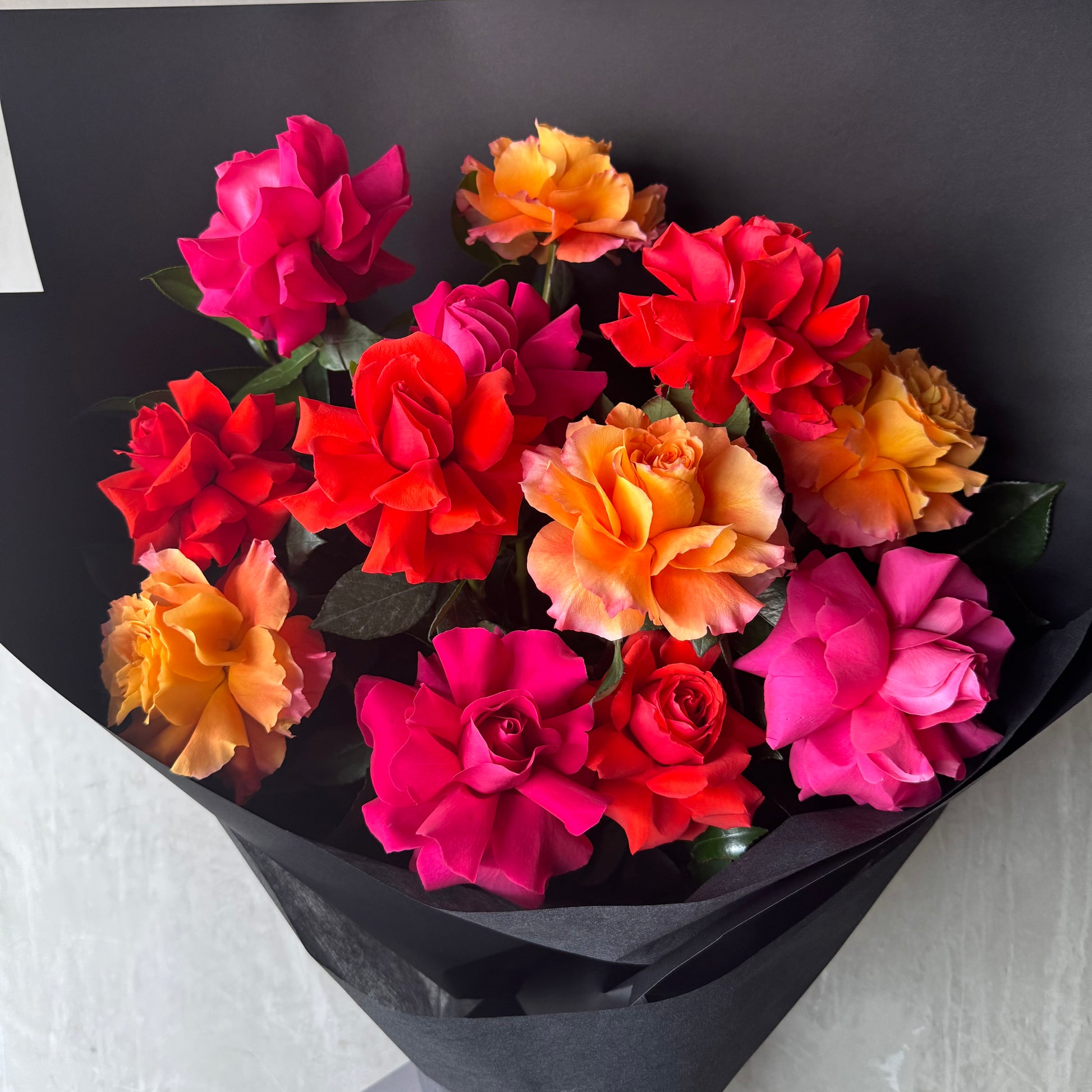 The Rose Trio Bouquet by FlowersonNortonSt features a stunning array of Colombian roses in vibrant pink, red, and orange hues, beautifully wrapped in black paper to highlight their rich colors and delicate petals.