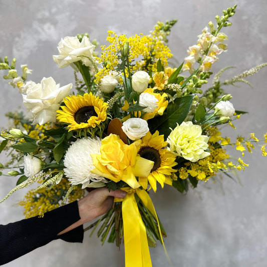 A hand showcases the vibrant Lemon Fizz bouquet from Flowers on Norton St, highlighting sunflowers, white roses, and yellow roses, complemented by lush greenery and small yellow chrysanthemums. A cheerful yellow ribbon wraps around the stems against a textured gray backdrop.