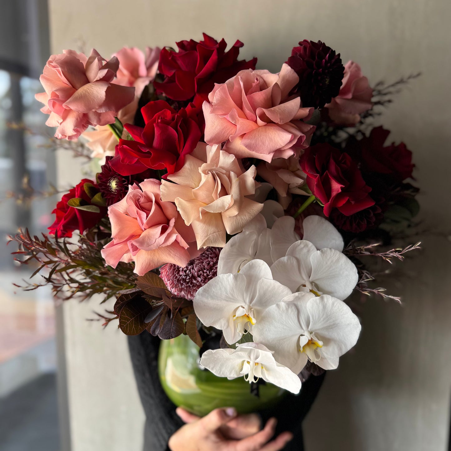 The Valentine's Dream Vase Arrangement by Flowers on Norton St showcases vibrant pink and Colombian roses, white phalaenopsis orchids, and assorted greenery in a round vase, set against a neutral background to accentuate its rich colors and textures.