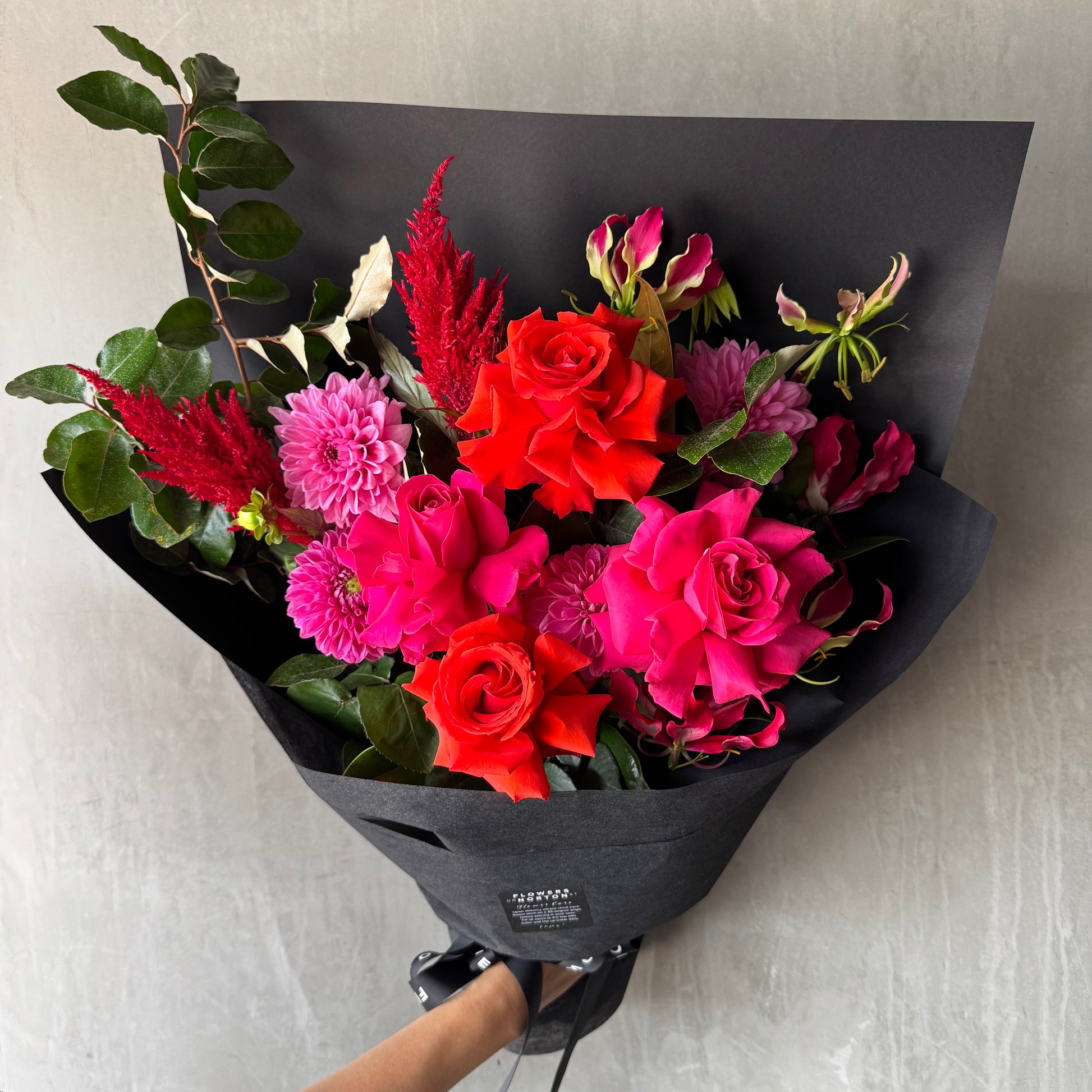 A bouquet featuring FlowersonNortonSt's Florist Choice - Red Nina Bouquet with vibrant Red Nina and pink roses, pink dahlias, and seasonal blooms with green leaves, elegantly wrapped in black paper against a gray background.