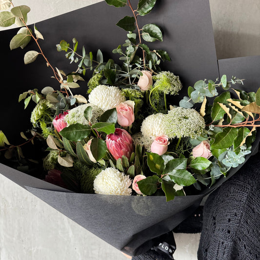 The Delicate Roses + Natives Bouquet by Flowers on Norton St is gracefully arranged with pink roses, white chrysanthemums, protea as native flowers, green foliage, and eucalyptus wrapped in black paper and elegantly placed on a gray surface.