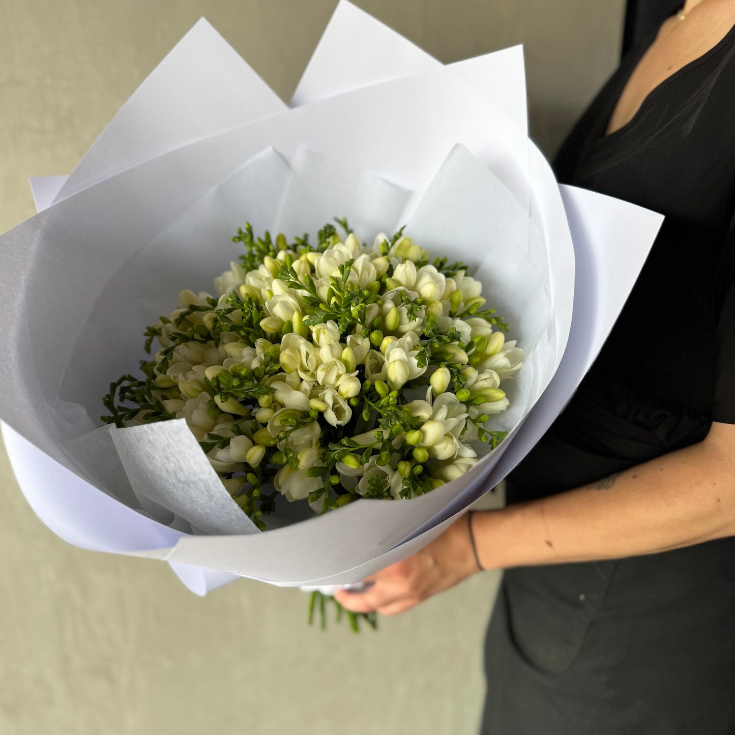 A person in a black outfit holds 'A Fantasy of Freesias Bouquet' by Flowers on Norton St, featuring fragrant freesias with delicate petals wrapped in white paper. The bouquet is neatly arranged, providing a gentle burst of color against the dark background.