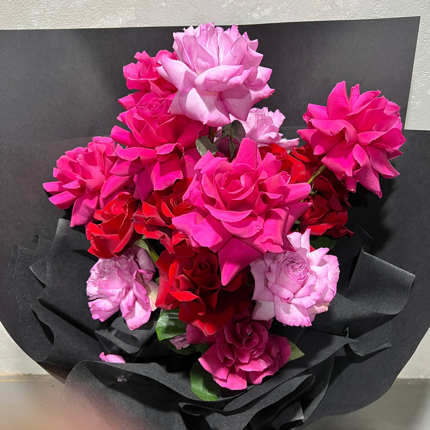 The Rose Trio Bouquet by FlowersonNortonSt showcases a vibrant mix of pink and red Colombian roses, elegantly wrapped in black paper against a neutral backdrop.