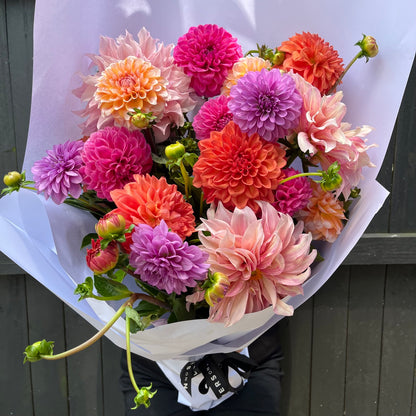 The Dahlia Delight bouquet from Flowers on Norton St features vibrant wedding flowers in shades of pink, orange, and purple, beautifully wrapped in white paper against a dark background. These fresh and lush blossoms showcase a variety of sizes and textures.