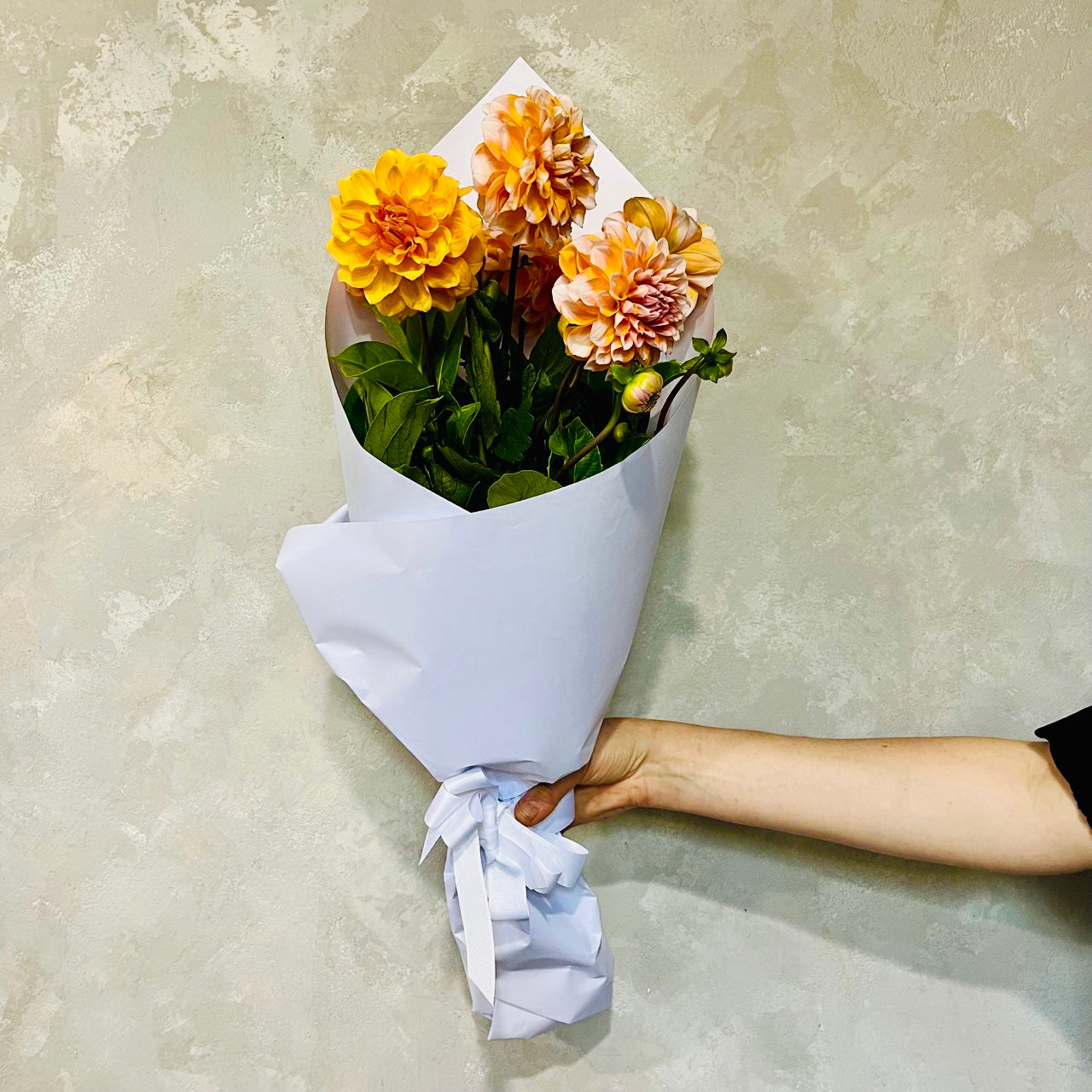 A hand holds a DAHLIA SPECIAL bouquet from Flowers on Norton St, featuring vibrant orange and yellow summer blooms wrapped in white paper, set against a textured greenish wall.