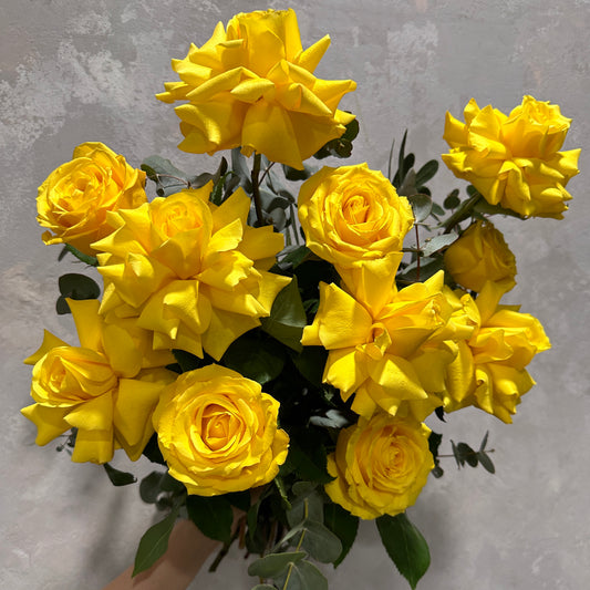 Experience the vibrant allure of Flowers on Norton St's Yellow Bud + Bloom Rose Bouquet, where radiant yellow roses contrast beautifully with lush green leaves against a textured gray background, offering a delightful sensory experience that captivates both sight and soul.